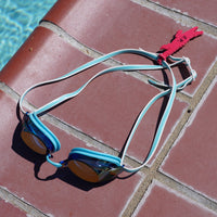 Dark pink alligator shaped Swim Loops goggle tag with name written on it attached to swim goggles on pool deck next to swimming pool. Make sure everyone knows those are your swim goggles. Get your Swim Loops goggle tags at www.swimloops.com.